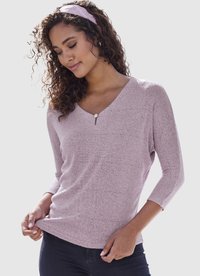 Frau mit lockigem Haar und einem rosa Stirnband, die ein strukturiertes, hellviolettes Langarmshirt mit V-Ausschnitt trägt und den Saum hält, vor einem einfarbigen Hintergrund.