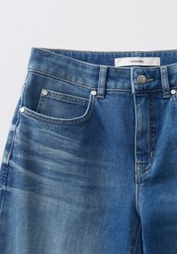 Jean en denim bleu avec poches avant, passants de ceinture, fermeture à bouton et une étiquette à l'intérieur de la taille portant la mention "un jour".