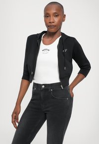Felpa nera corta con zip e logo bianco, indossata sopra una canotta bianca con testo stampato. Abbinata a jeans skinny di denim scuro.