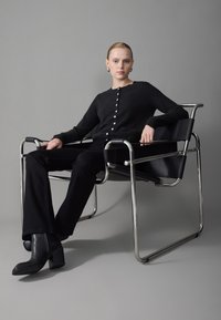 Black button-up cardigan made of soft fabric, worn with black flared pants and textured black heeled boots, seated in a chrome chair.