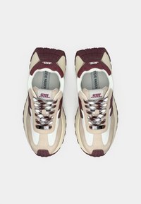 Baskets composées de matériaux beige et bordeaux, semelles texturées, accents blancs, lacets imprimés, et un logo bien visible sur la languette.