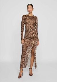 Kvinna i en långärmad maxiklänning med brun leopardtryck och en slits fram, bärande svarta högklackade sandaler, som står framför en enfärgad bakgrund.