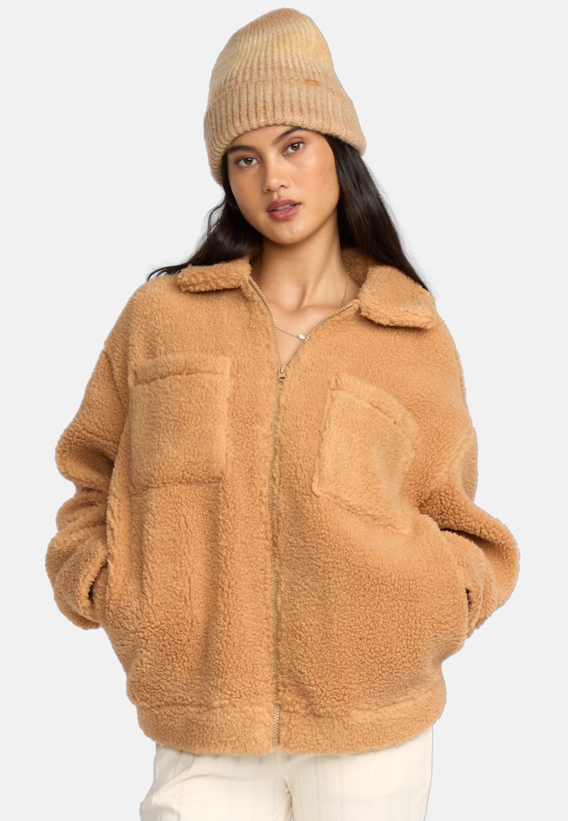 Giacca in pile color sabbia con due tasche frontali, chiusura con zip e colletto. Indossata con un berretto rigato coordinato. Tessuto morbido, vestibilità oversize.