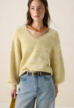 Femme portant un pull à col en V marbré jaune, un jean taille haute bleu clair avec une ceinture marron, un collier de perles ambrées, tenant une poignée de sac moutarde.