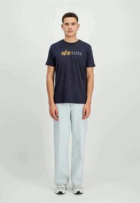 Άνδρας που στέκεται φορώντας ένα ναυτικό μπλουζάκι Alpha Industries, ανοιχτό μπλε τζιν και άσπρα sneakers σε απλό φωτεινό φόντο.