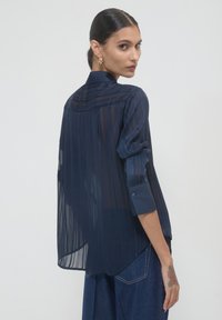 Blusa trasparente blu navy con strisce verticali, colletto alto e polsini con bottoni. Il tessuto sembra leggero con una silhouette fluida.