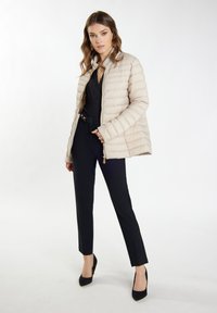 Giacca imbottita beige chiaro con zip frontale, cuciture orizzontali e colletto alto, abbinata a pantaloni neri su misura e tacchi neri.