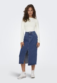 Denim midi skirt with a front slit, high waist, and classic five-pocket design. Paired with a fitted white long-sleeve top and white sneakers.