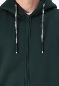 Zöld cipzáros kapucnis felső, puha anyagból készült. Felszerelt egy zsinóros kapucnival, fekete és fehér részletekkel, valamint elöl cipzárral. Egyszerű dizájn.