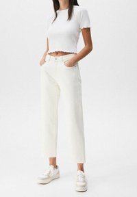Top corto bianco a costine con maniche corte a campana, abbinato a jeans crema a vestibilità rilassata e sneakers bianche con accenti beige.