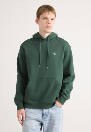 Groene capuchontrui van een katoenmix, met een voorzak, geribde manchetten en een klein gouden CK-logo. Gecombineerd met lichtblauwe spijkerbroeken.