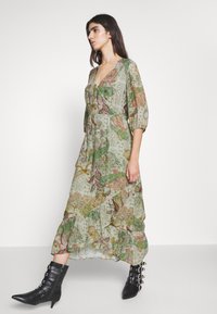 Grünes Blumenkleid im Maxistil mit Puffärmeln, Rüschensaum und V-Ausschnitt. Kombiniert mit schwarzen Stiefeletten mit silbernen Schnallen.
