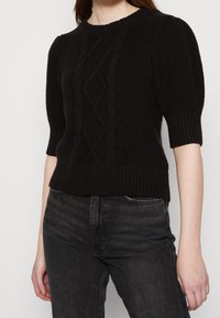 Pull en tricot noir à manches ballon courtes, avec un design texturé en câble et un col et un ourlet côtelés. Porté avec un jean noir.