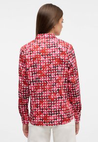 Langärmliges Hemd aus rot-rosa Stoff mit einem kreisförmigen Punktmuster, ausgestattet mit einem Kragen und geknöpften Manschetten. Glatte Textur, taillierte Passform.