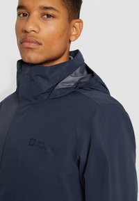 Veste imperméable marine avec col haut et capuche doublée en maille. Présente un logo sur la poitrine, avec une texture extérieure lisse et mate.