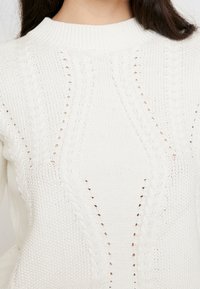 Knitted white sweater featuring a ribbed neckline, textured patterns, and open stitches creating a breathable design.