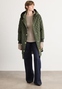 Geverfde donkere groene parka met een capuchon, gouden drukkers en een rits aan de voorkant. Draag het over een beige gebreide trui en donkere flared jeans. Binnenomgeving.