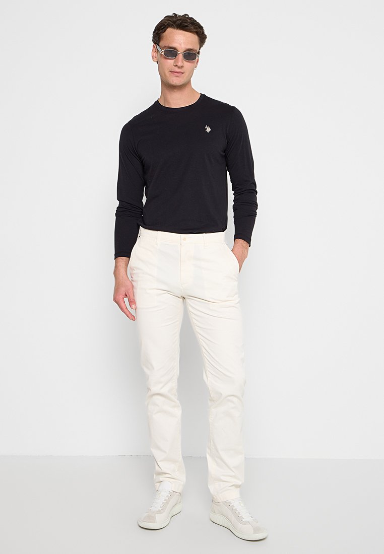 Homme portant une chemise noire à manches longues, un pantalon blanc, des baskets blanches et des lunettes de soleil foncées, se tenant avec une main dans la poche sur fond blanc.