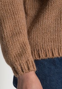 Gebreide trui, lichtbruin, met ribbels aan de mouwen en onderkant, zachte textuur, gedeeltelijk te zien op een arm tegen een donkere jeans.
