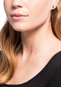 Ung kvinna med ljus hy och långt ljusbrunt hår, som bär blåa ädelstens-studörhängen och en svart ribbad topp.