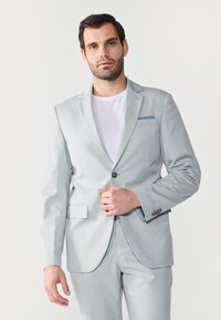 Costum gri deschis, cu un sacou cu un singur nasture, rever în formă de V, buzunare laterale și un batistă albastră în contrast. Material neted.