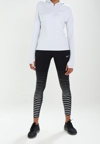 Maglione grigio chiaro a maniche lunghe con zip e cappuccio, abbinato a leggings a righe bianche e nere. Presenta il logo Nike e scarpe da ginnastica nere.