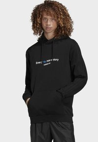 Sort hættetrøje med en frontlomme, der har hvid og blå tekst "Alle har en historie" og adidas-logo. Tekstureret stof med ribbede manchetter.