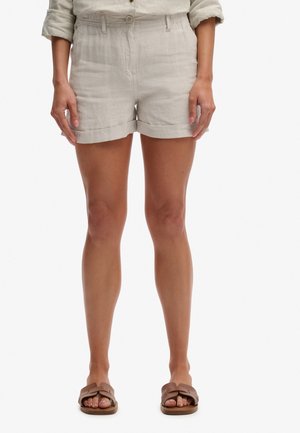 Donna che indossa pantaloncini beige con orlo arrotolato, una camicia beige a maniche lunghe con le maniche arrotolate e sandali marroni a ciabatta, in piedi dritta.