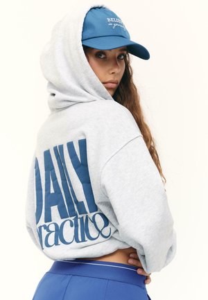 Femme portant un sweat à capuche gris avec le texte "DAILY practice" et une casquette bleue sur laquelle est inscrit "BELIEVE in yourself", regardant par-dessus son épaule.