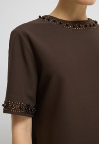 Brązowa bluzka z krótkim rękawem i okrągłym dekoltem, ozdobiona ciemnymi cekinami i koralikami wzdłuż kołnierza oraz brzegów rękawów.