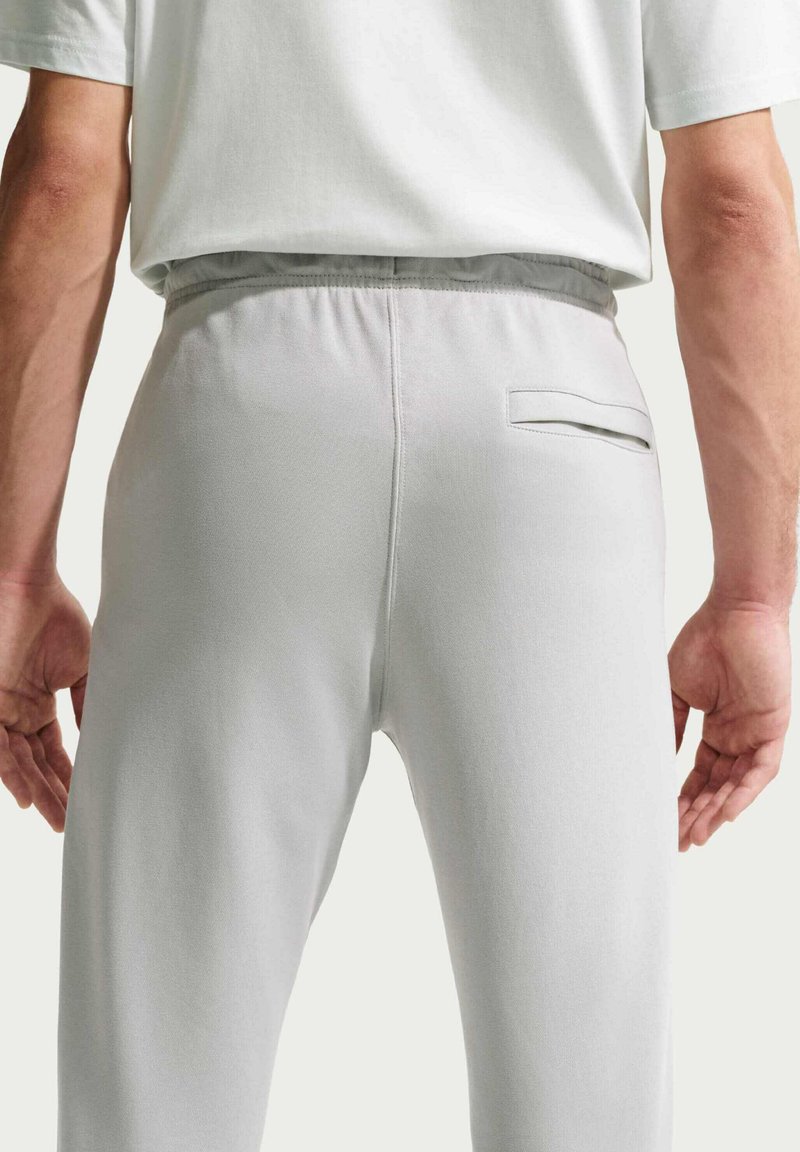 Hombre con pantalones gris claro con cintura elástica y un bolsillo de ojal en la parte trasera, de pie con los brazos relajados a los lados.