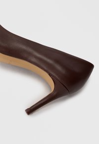 Chaussure à talon haute en cuir marron avec un bout arrondi, texture lisse et un talon aiguille mince. Design simple, sans motifs ni accents visibles.