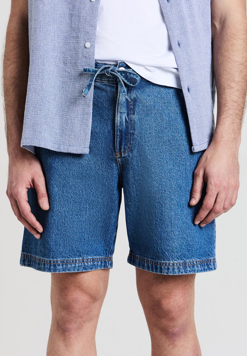 Homme portant un short en denim bleu à mi-thigh avec une taille nouée, un t-shirt blanc et une chemise à boutons ouverte de couleur bleu clair.