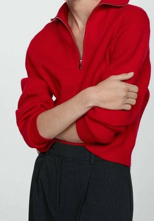 Maglione rosso a coste con zip a quartino e collo alto. Indossato con pantaloni scuri, con polsini larghi e vestibilità comoda.