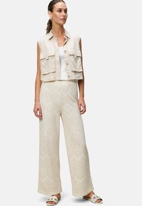 Gilet beige court avec deux poches avant, associé à un pantalon large en dentelle crème présentant des motifs intricats. Porté avec des sandales blanches.