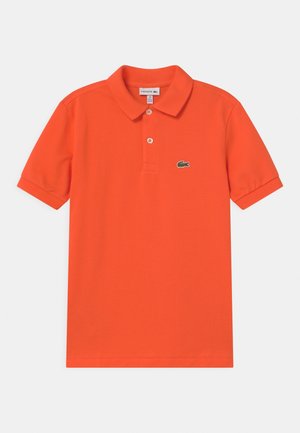 Pomarańczowa koszulka polo z bawełny, z krótkim rękawem, klasycznym kołnierzykiem, zapięciem na trzy guziki oraz zielonym logo krokodyla na klatce piersiowej.