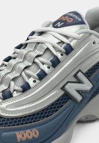 Silver and navy sneaker with mesh upper, textured lacing, rubber sole, and prominent 'N' logo. Features contrasting accents and stitching details.