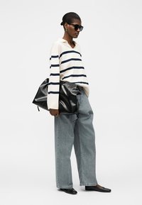 Γυναίκα που φοράει μαύρα γυαλιά ηλίου, λευκό πουλόβερ με μαύρες ρίγες, άνετα μπλε τζιν, μαύρα μπαλαρίνες και κρατά μια μεγάλη μαύρη δερμάτινη τσάντα.