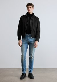 Čierna bomberová bunda so zapínaním na zips, nosená cez čierny rolák, doplnená o vybledlé svetlomodré džínsy a čierne tenisky.