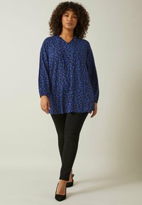 Blå leopardmönstrad tunika med långa ärmar, V-ringning och en avslappnad passform, matchad med svarta skinny jeans och svarta klackskor.