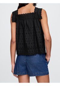 Kvinna i en ärmlös svart broderad topp med scallopdetaljer och blå denimshorts, stående med en hand i fickan.