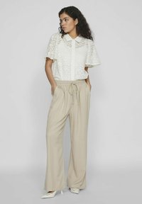Blouse en dentelle blanche avec des manches courtes et larges, dotée d'une fermeture à boutons, associée à un pantalon beige, ample, avec cordon de serrage et des chaussures blanches à pointe.