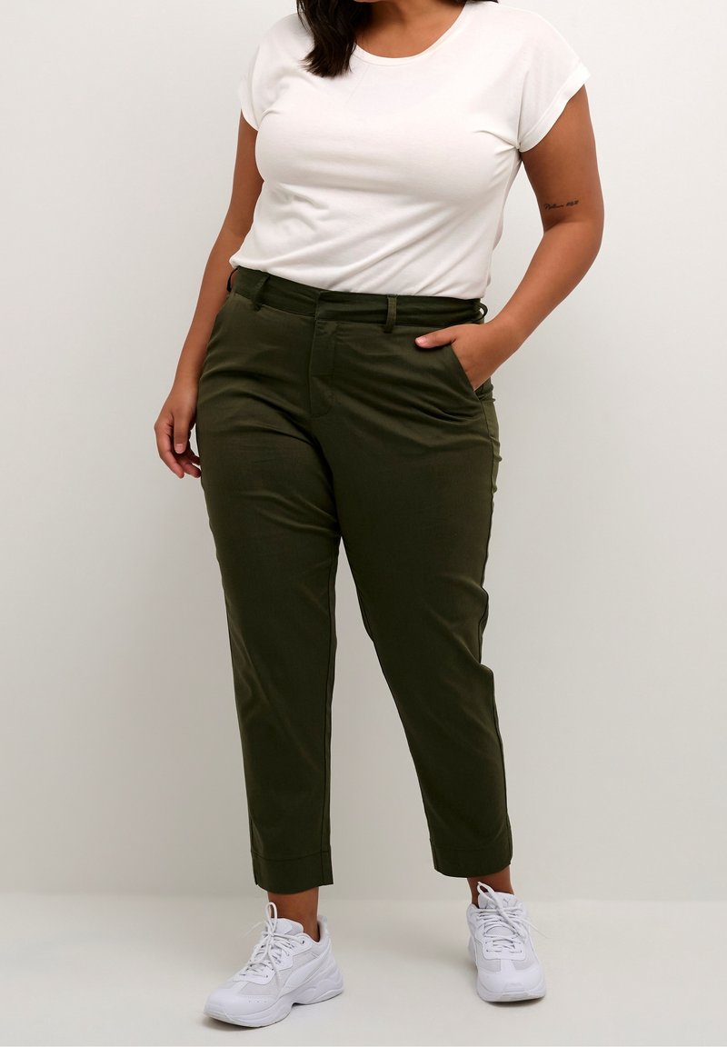 Femme portant un t-shirt blanc à manches courtes, un pantalon olive longueur cheville, et des baskets blanches, debout avec une main dans la poche.