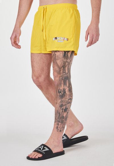 Costumi da bagno gialli con logo Moschino, vita con cordino e superficie liscia. Sandali neri con fascia logata.