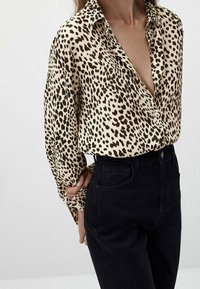 Beige knap-up skjorte med brune leopardpletter, der har en dyb V-udskæring og lange, let puffede ærmer, parret med højtaljede sorte jeans.