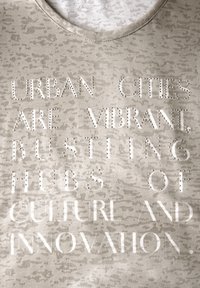 Camiseta gris claro con un patrón texturizado. Presenta pedrería en relieve formando el texto: "LAS CIUDADES URBANAS SON CENTROS VIBRANTES Y BULICIOSOS DE CULTURA E INNOVACIÓN."