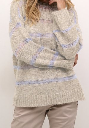 Femme portant un pull en maille beige avec des rayures horizontales bleu clair et un pantalon de couleur claire, posant avec une main près du visage.