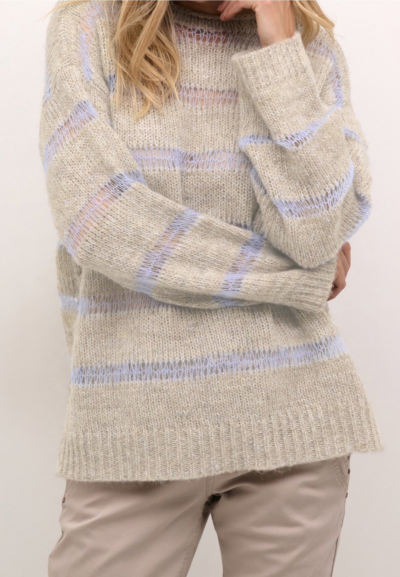 Femme portant un pull en maille beige avec des rayures horizontales bleu clair et un pantalon de couleur claire, posant avec une main près du visage.