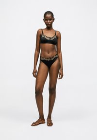 Femme aux cheveux tressés portant un maillot de bain deux-pièces noir avec un bord beige ondulé, debout pieds nus devant un fond blanc uni.