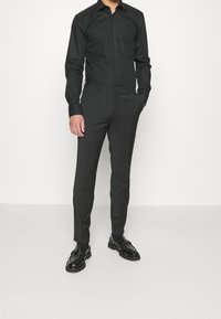 Man in een zwart overhemd met knopen, zwarte op maat gemaakte broek en zwarte gepolijste nette schoenen, staand met één hand in zijn zak.